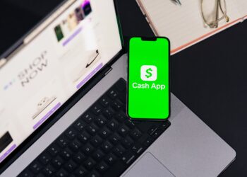 Stock photo of a smartphone displaying the Cash App logo on a green screen, resting on a laptop keyboard with an online shopping page open in the background, alongside a notebook, pen, and glasses.